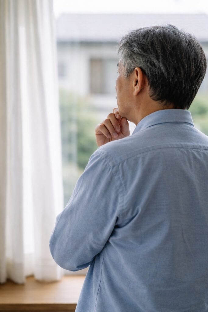 窓辺で立ち止まり考え込む60代男性の後ろ姿